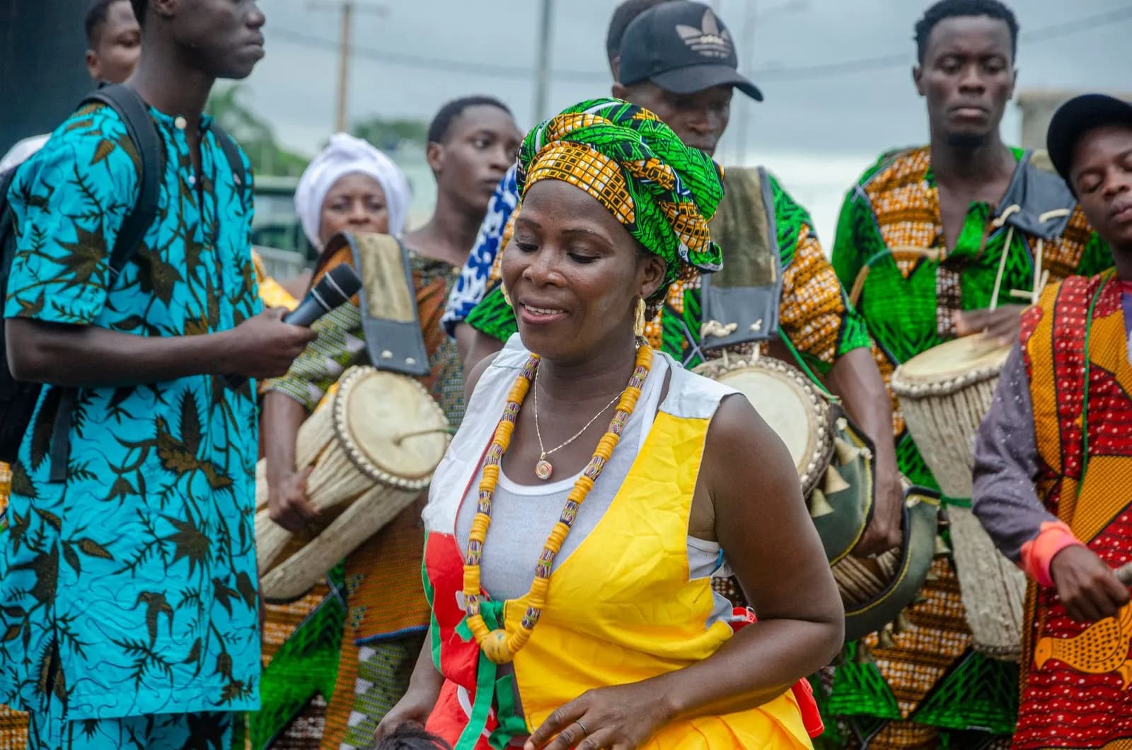 A street celebration in Aného, Togo: a dancer in yellow with bead necklaces, drummers in bright kente behind her